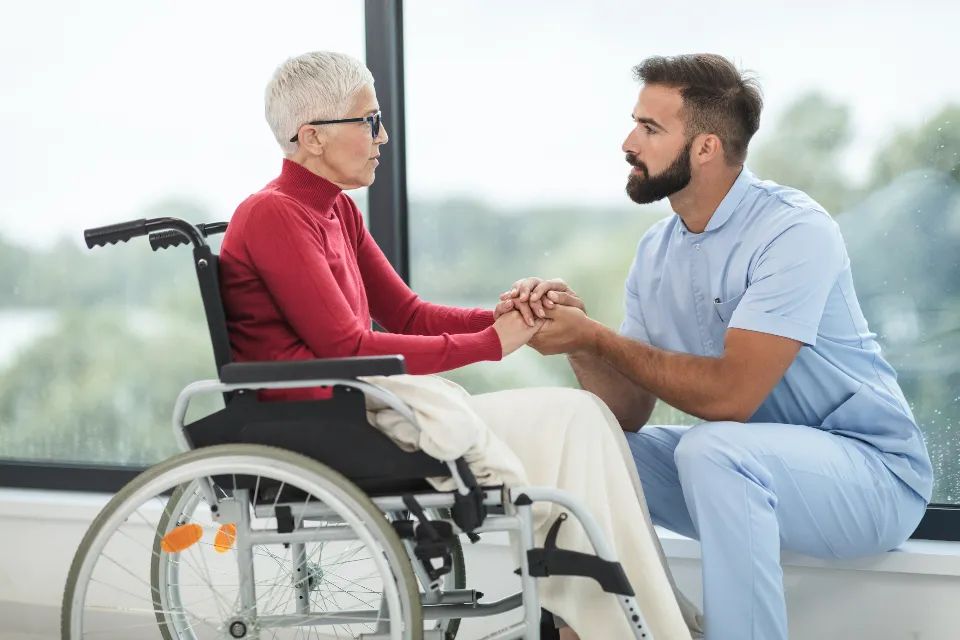 A caregiver in blue scrubs holding the hand of a senior in a wheelchair