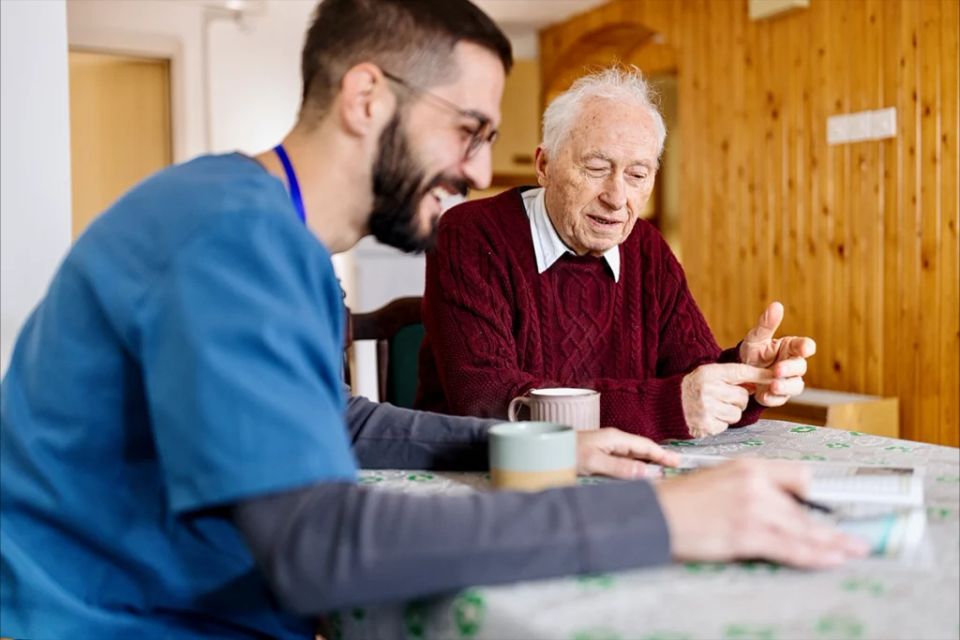 A nurse in blue scrubs in conversation with a senior during a care assessment at home