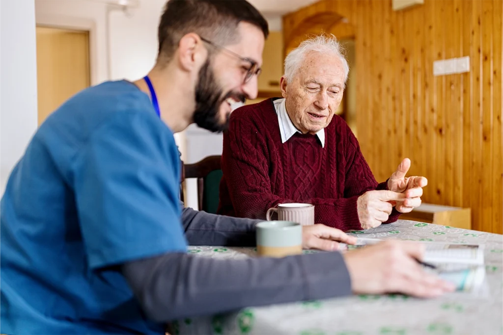 A nurse in blue scrubs in conversation with a senior during a care assessment at home
