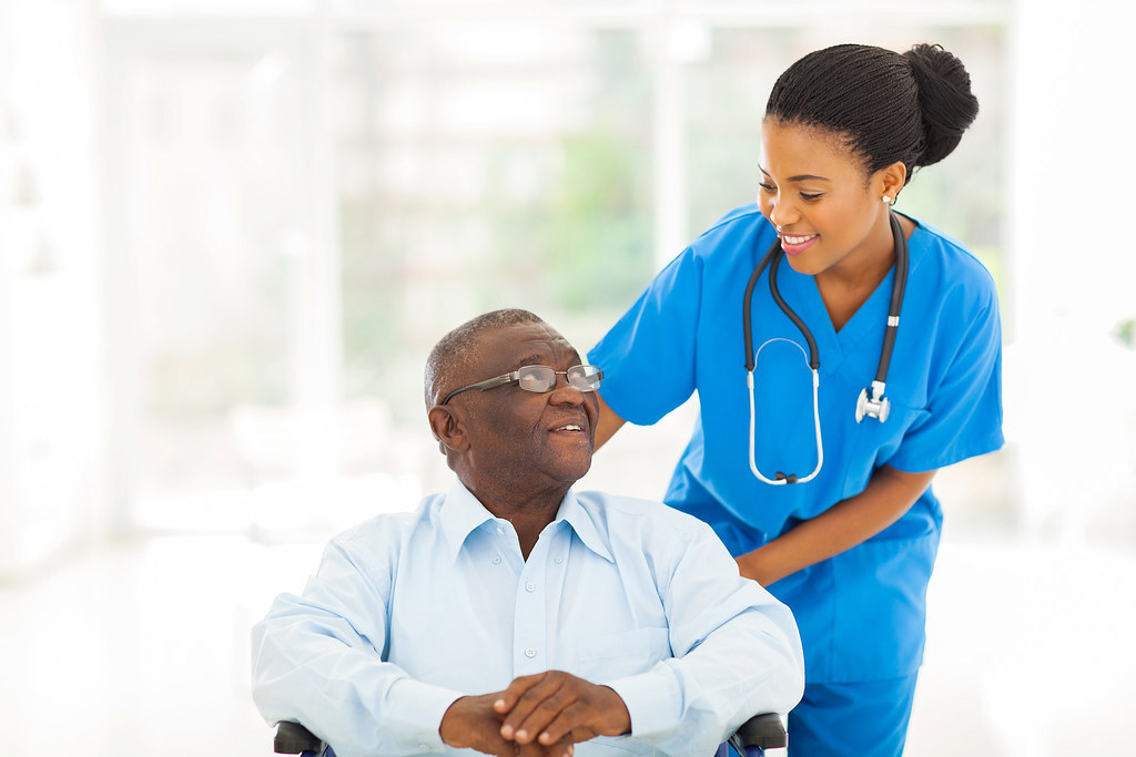 A nurse in blue scrubs assisting an older man recovering in a wheelchair
